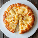 Ein goldbrauner Birnenkuchen auf einer weißen Platte, geschnitten mit frischen Birnenscheiben und Zimt bestreut, umgeben von einem rustikalen Holztisch.