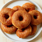 Goldbraune französische Crullers auf einem weißen Teller; luftige Innenstruktur sichtbar, knusprige äußere Kruste, leicht bestäubt mit Puderzucker; Teller auf hellem Holztisch, unscharfer Hintergrund, natürliche Beleuchtung.