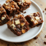 Frischkäse-Brownies auf einer warmbraunen Holzplatte, zwei Stücke mit dunkler Schokoladenbasis und cremiger Frischkäse-Swirl, strukturierte Oberfläche, sanft glänzend, rustikales Setting.