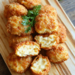 Ein Teller mit goldbraunen Brokkoli-Käse-Nuggets, knusprig und saftig, präsentiert auf einem hellen Holzteller. Im Hintergrund leicht verschwommene grüne Brokkoliröschen, rustikale, einladende Präsentation, dampfige Note, appetitlich und frisch.