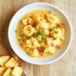 Eine cremige Blumenkohl-Curry-Sauce mit goldbraunen Ofen-Blumenkohlröschen in einer weißen Schale. Frischer Koriander oben drauf, heller Holztisch als Hintergrund, warme natürliche Beleuchtung, Nahaufnahme mit sichtbarer cremiger Textur.