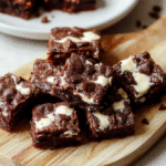 Nahaufnahme von Frischkäse-Brownies Deluxe: quadratische Brownies mit goldbrauner Oberfläche und cremiger Käse-Schicht, dunkle Schokoladenstücke an den Rändern, angerichtet auf heller Holzplatte, sanfte Schatten