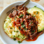 Close-up eines Burrito Bowls: gebräunte Hackfleischportionen, gelber Reis, schwarze Bohnen, Mais, rote Paprika, Avocado-Scheiben, Käse, Salsa und frische Korianderblätter auf einem Teller; farbenfroh, cremig, rustikal.