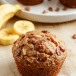 Goldbraun gebackene Protein-Bananen-Muffins in Papierförmchen auf einem hellen Holztisch, sichtbare Bananenstücke, saftige Innenstruktur und leichte Krustenränder.