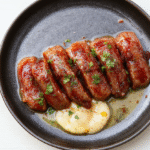 Eine Pfanne mit Würziger Wurst Reis Pfanne zeigt goldbraune Würstchen, locker gekochten Reis, rote Paprikastücke und Zwiebelringe. Garnitur mit Petersilie, serviert auf einem hellen Holztisch. Die Farben Goldbraun, Gelb und Rot dominieren, Texturen von gebräunter Wurst und körnigem Reis wirken einladend. Nahaufnahme, warme Lichtstimmung, appetitlich und rustikal.