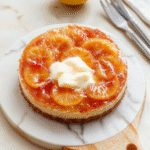 Eine Zitronen-Joghurt-Torte auf einem weißen Teller, glatte Oberfläche, dekoriert mit Zitronenzesten und einem Spritzer Zitronensaft; heller neutraler Hintergrund, natürliche Beleuchtung, sanfte Schatten.